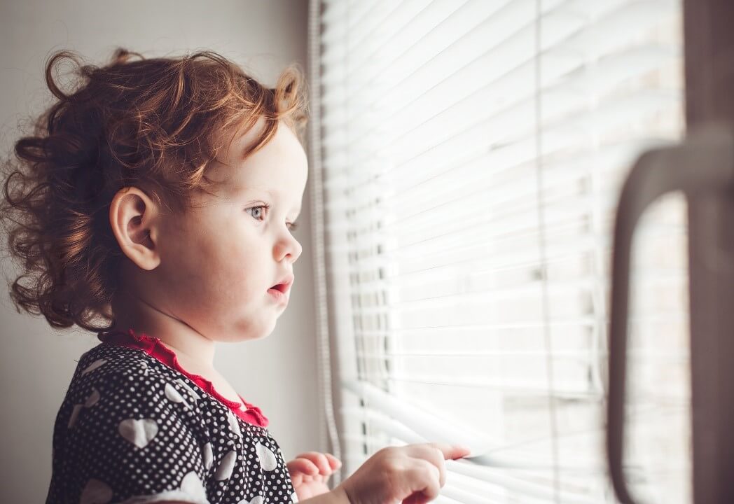 small child looking out through a window with blinds